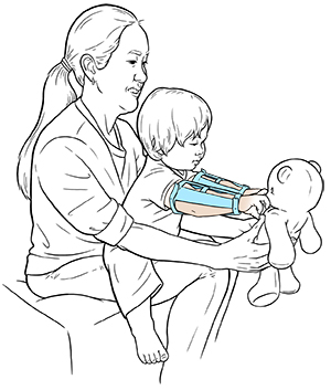 Toddler with arm restraints playing with teddy bear while sitting in woman’s lap.