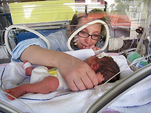 Woman looking at baby in incubator, with arm inside giving light touch to baby.