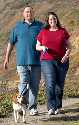 Man and woman outdoors walking dog.