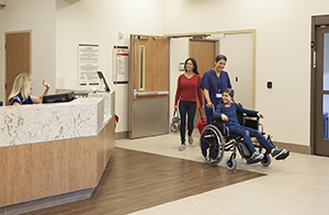 Healthcare provider pushing girl in wheelchair towards hospital exit with woman walking alongside.