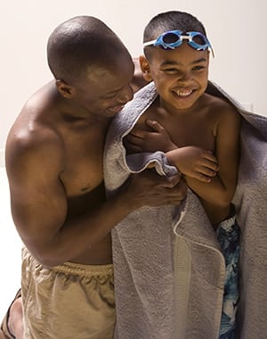 Man holding preschool-age boy in towel near swimming pool.