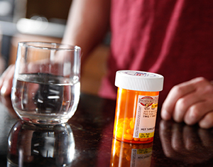 Closeup of pill bottle and glass of water with person in background.